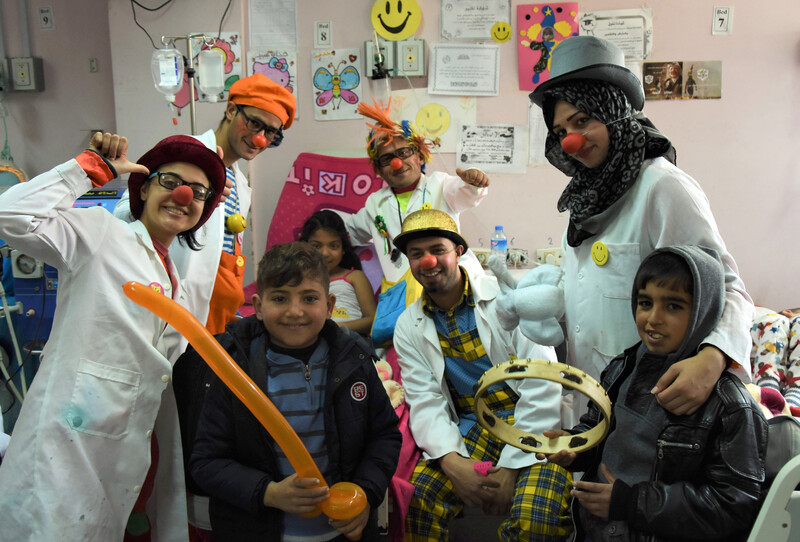 Men and women in silly hats and wigs wearing red foam noses pose with children in hospital room