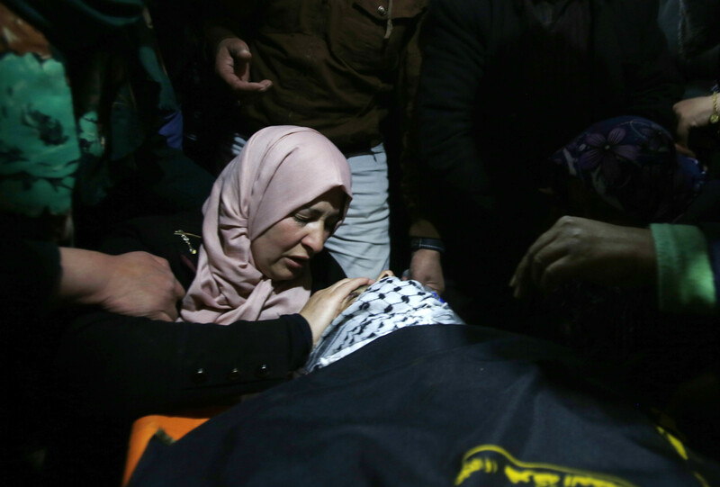 Crying woman places hand on forehead of body wrapped in black flag and checkered scarf