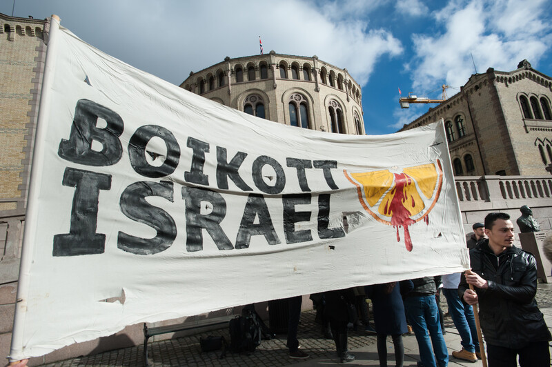 A large banner reading Boikott Israel with an image of a blood-splattered orange slice is held in front of a building