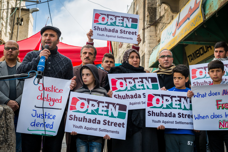 Man speaks into microphone accompanied by men, women and children holding signs reading Open Shuhada Street