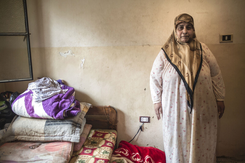 Middle-aged woman wearing headscarf and cotton dress stands next to bed in sparely furnished room