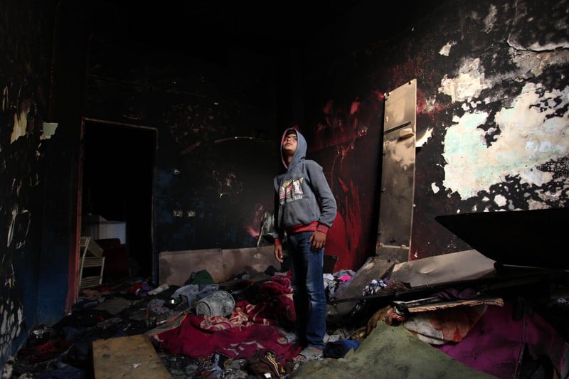 A boy stands in the middle of a room with charred walls and debris