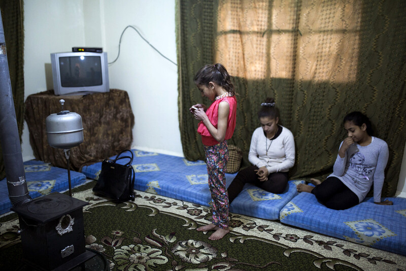 Girl stands in middle of room looking at device as two older girls sit on mats on floor in living room