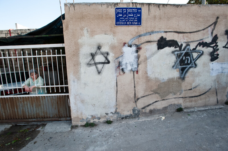 Woman stands behind bars next to walls spray-painted with Stars of David