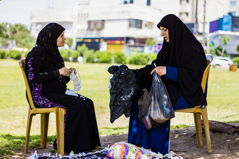 Women sit on chairs while crocheting and holding up plastic