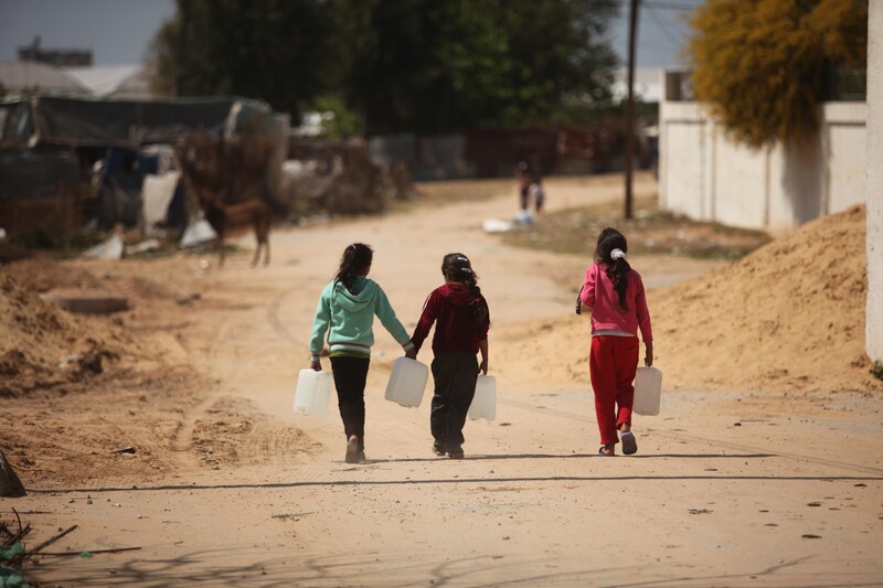 Three children carry large containers of water as they walk along a dirt road