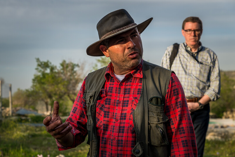 Man wearing wide-brimmed hat and vest over flannel shirt gestures with his hand