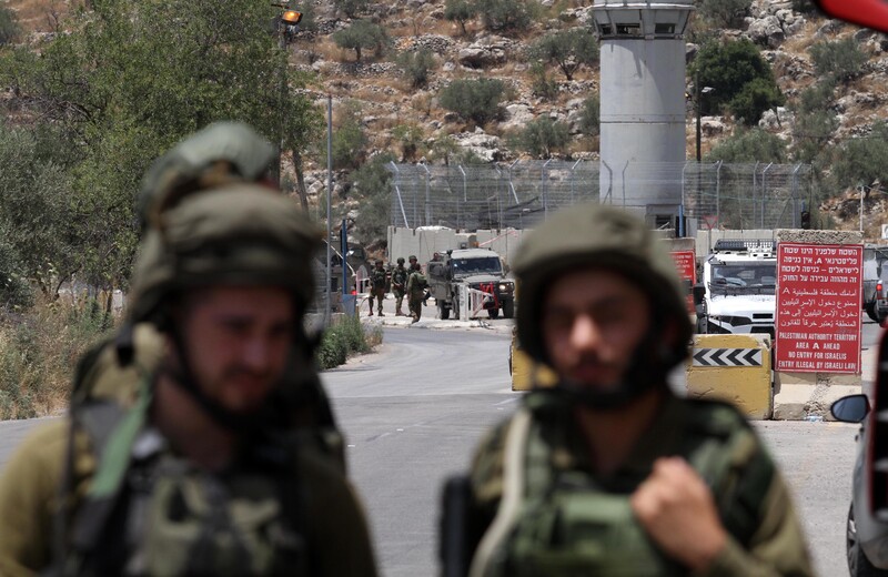 Israeli soldiers stand in foreground with military checkpoint watchtower and crowd of people in background