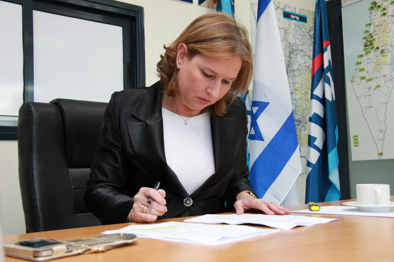 Tzipi Livni sits at desk looking at paper with an Israeli flag hanging behind her