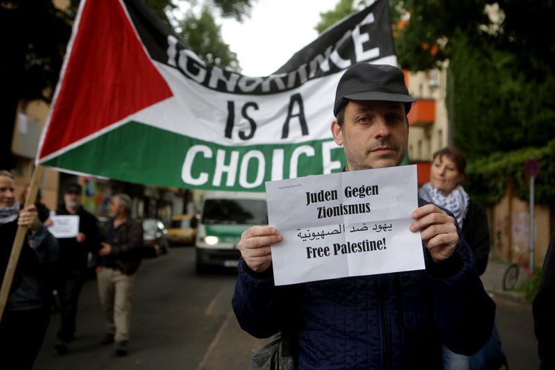 Man carries sign reading Jews against Zionism in German and Arabic and Free Palestine in English