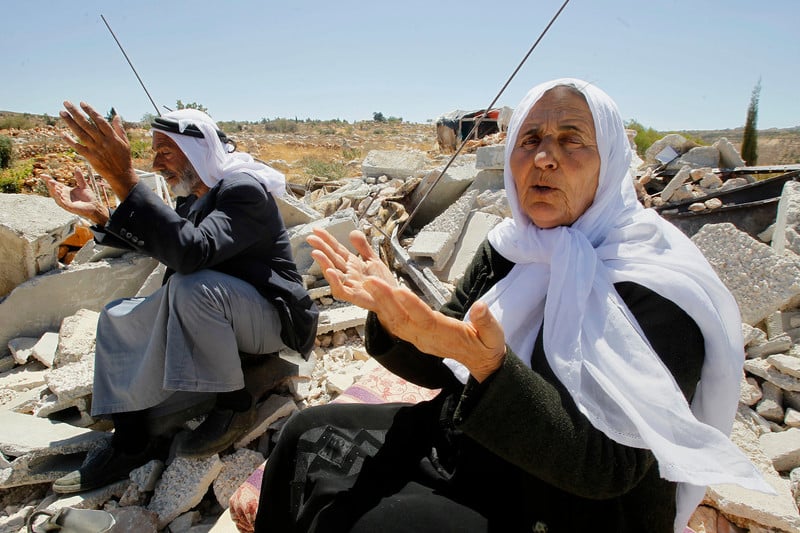 Elderly couple gesture with their hands while sitting on rubble of destroyed home