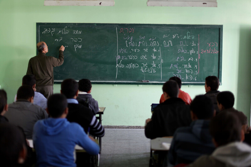 Man writes words in Hebrew on chalkboard in front of students sitting at desks