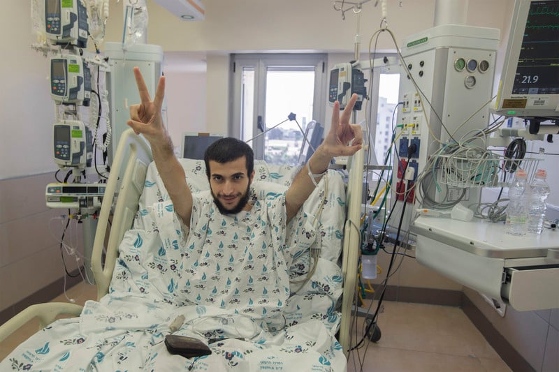 Smiling man in hospital bed raises his arms with his fingers in V for victory sign