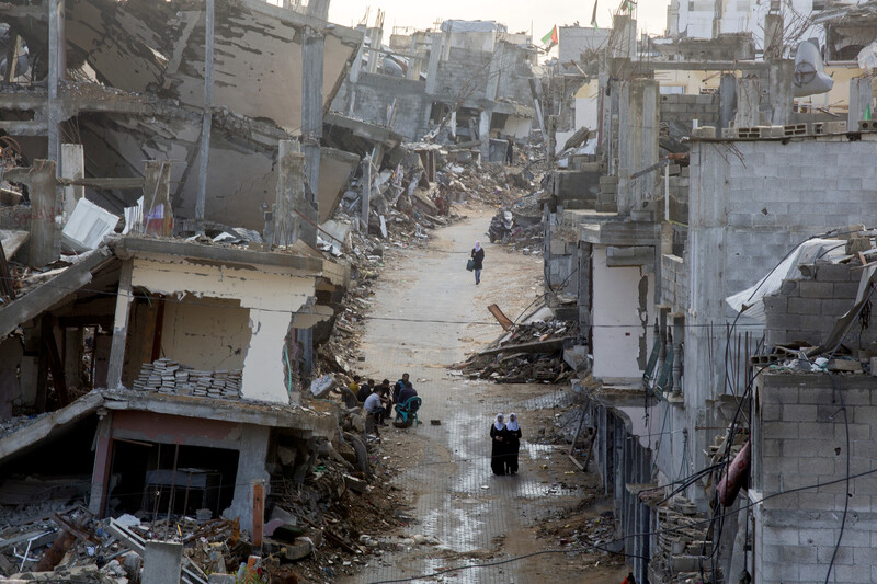 View of people walking through muddy road in between bombed-out buildings