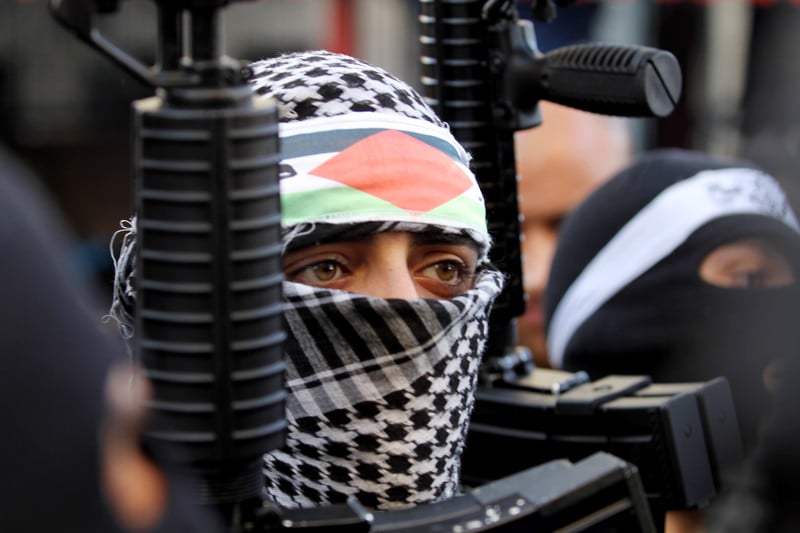 Young man wearing checkered scarf around his head and face holds rifle