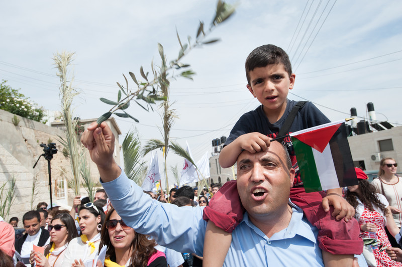 Man waving olive branch carries boy holding Palestine flag on his shoulders during Palm Sunday procession