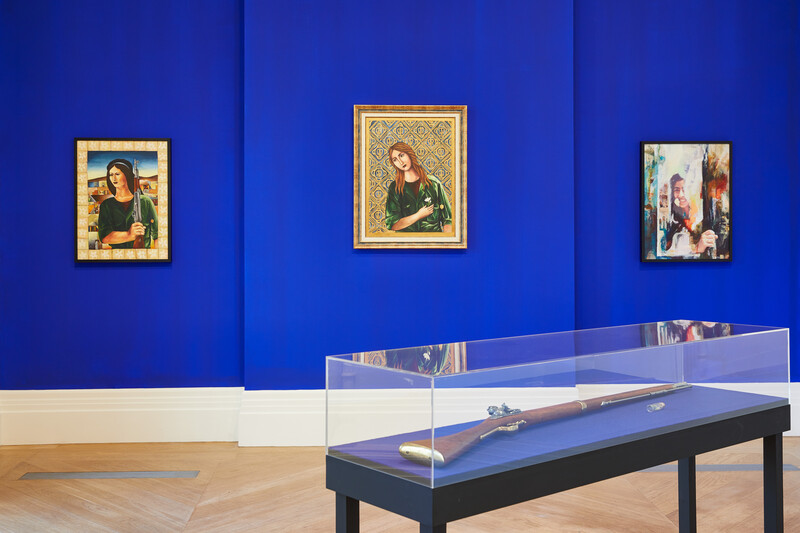 Installation view of three works on canvas on wall with vitrine containing rifle in foreground