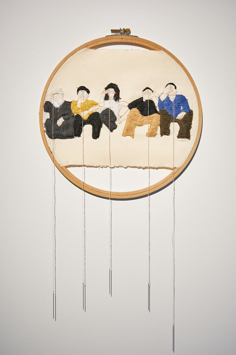 Embroidered panel framed by embroidery hoop shows five seated figures with a threaded needle hanging from each