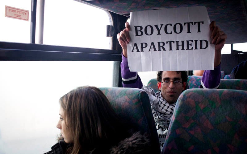 Basil al-Araj sits on bus behind Israeli woman while holding up a sign reading Boycott Apartheid