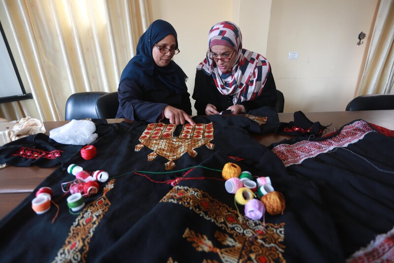 Two women sitting at table lean in and look at embroidered panel on dress