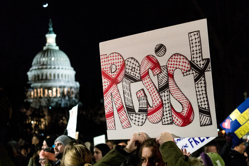 Woman holds sign reading Resist done in pattern of checkered Palestinian scarf in front of Capitol Hill dome during nighttime protest
