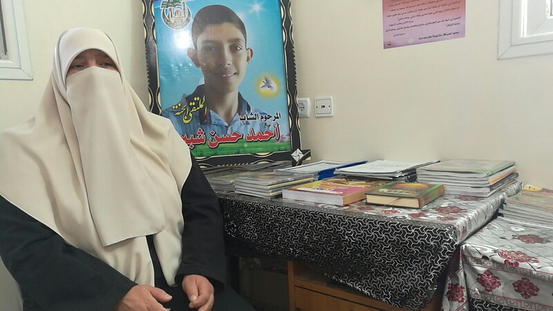 Woman with covered face sits next to table and poster of her smiling teenage son