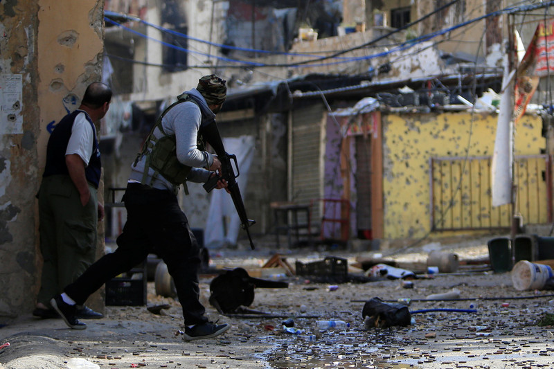 Man carrying rifle and unarmed man look around a corner in a bullet-strewn, narrow street in Ein al-Hilweh