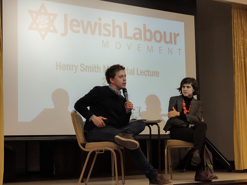 A man on stage talks into a microphone while a woman watches