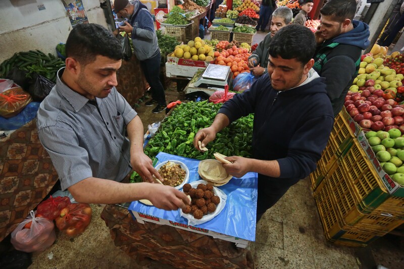 Two men stand at table making falafel sandwiches in middle of produce shop