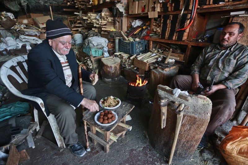 Elderly man sits on chair next to table holding small dishes of food in shop filled with wood