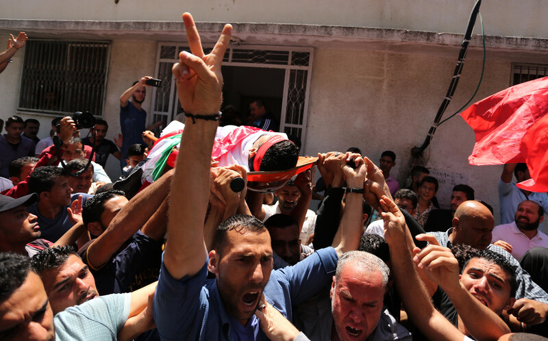 Man makes a V symbol with his right hand while Muhammad Bakr's shrouded body is carried on a stretcher in a crowd of people