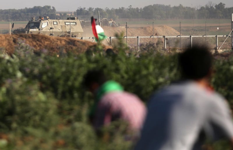 Photo shows backs of youths crouching in agricultural field, one holding a Palestinian flag, with an Israeli military vehicle in the background