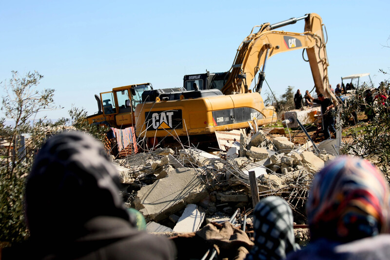 Photo shows the backs of women's heads as they watch a Caterpillar machine being used to raze a home