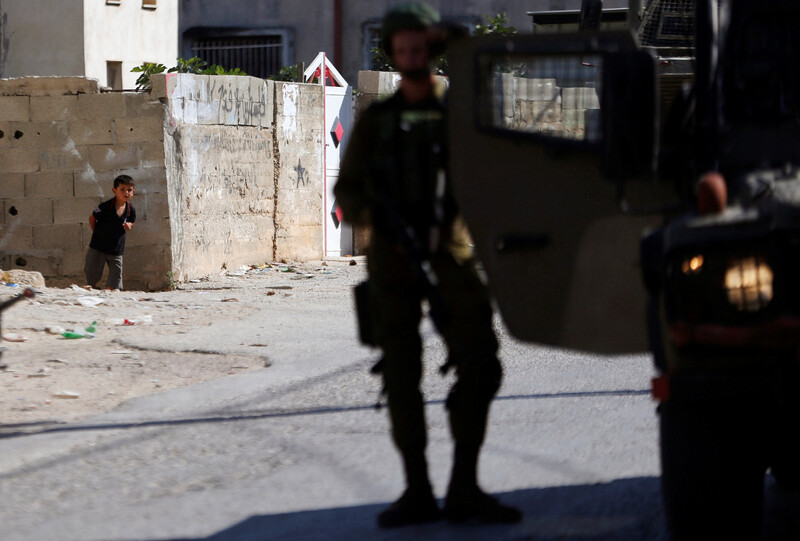 A small Palestinian boy peers behind a wall as heavily armed soldiers stand in village street