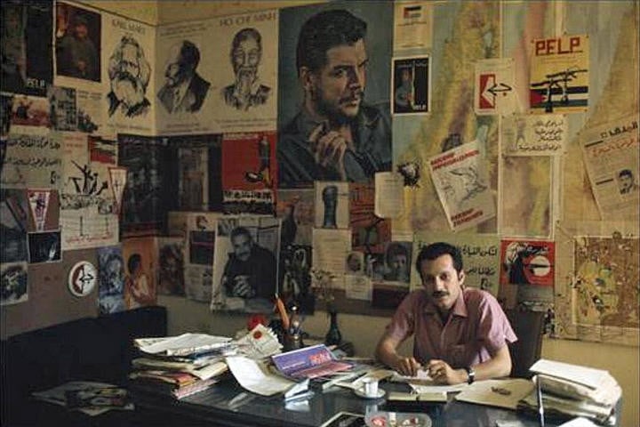 Ghassan Kanafani at his desk, behind him are political posters and fliers 