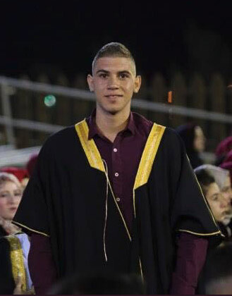 Photo shows smiling young man wearing graduation ceremony gown from waist up