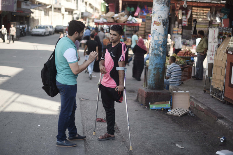 Two young men, one using arm braces, speak to each other in a marketplace