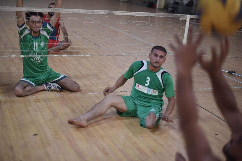 Young man wearing sports jersey sits on the ground behind a volley ball net, watching for the ball to be served