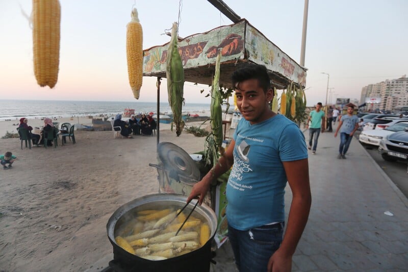 Teenager seen from waist up cooks corn in a vat in front of a stand on Gaza's corniche