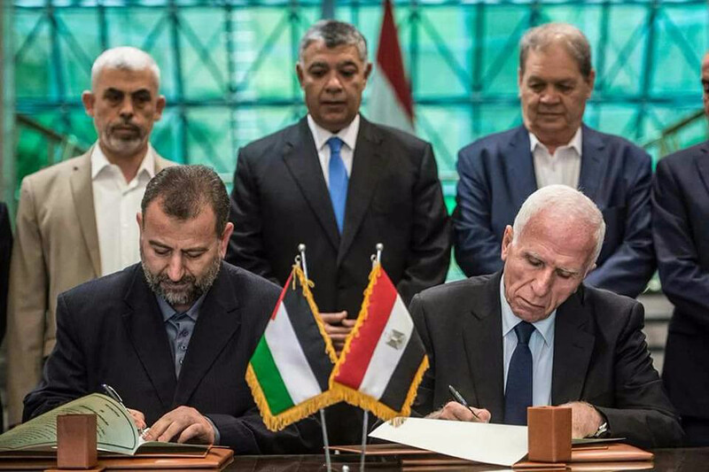Two men sitting at a desk sign documents as three men standing behind them look on