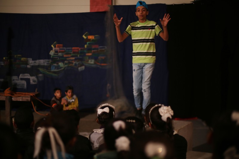 A boy wearing a T-shirt, jeans and a baseball camp puts his arms up while he stands on stage in front of a group of schoolgirls
