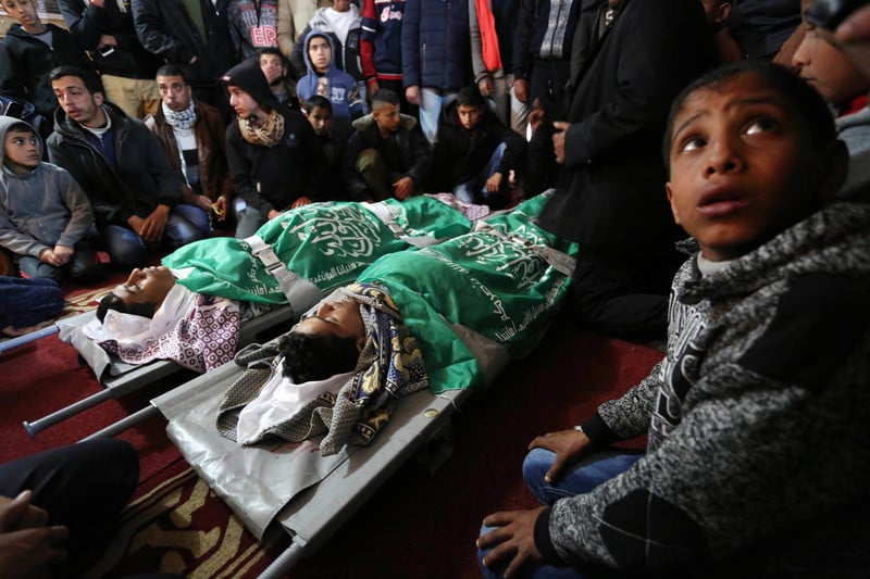 The shrouded bodies of two boys, wrapped in green flags, lay on stretchers while surrounded by mourners