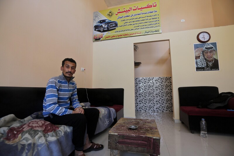 Young man sits on couch in sparely furnished room with a poster of Yasser Arafat on the wall