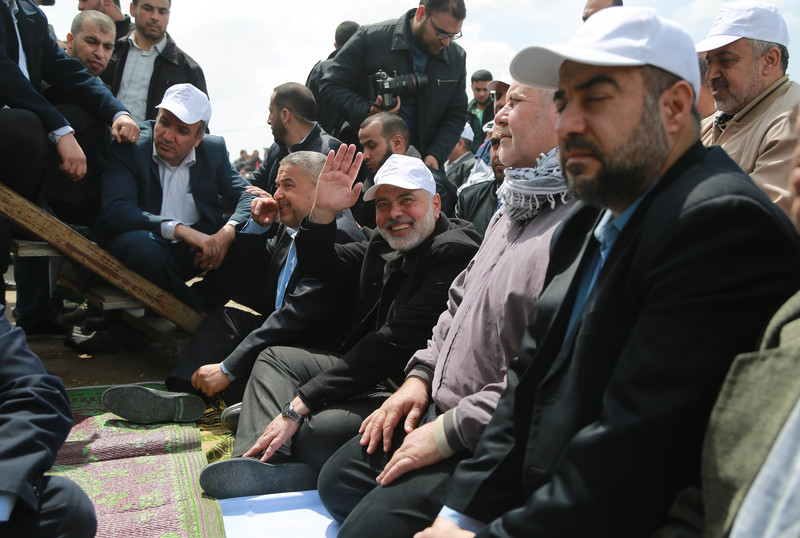 Ismail Haniyeh, wearing white baseball cap, sits on ground and waves towards camera