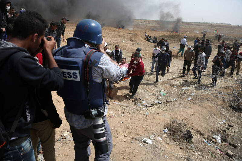 Photographer wearing flak jacket marked as PRESS and wearing a blue helmet is seen from behind while taking photos of demonstrators