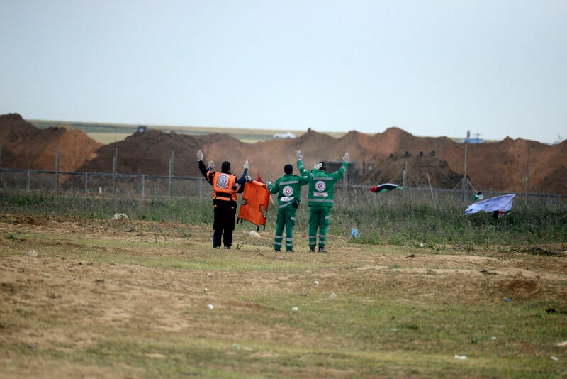 Three medics carrying a folded up stretcher put their arms in the air in front of Gaza-Israel boundary fence