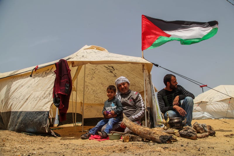 Two men and a small boy sit outside of a cloth tent with a Palestinian flag raised above it