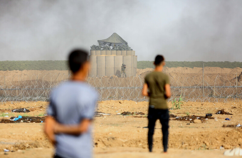The backs of two standing youth are seen in foreground of photo with Israeli military installation behind barbed wire and fencing in background