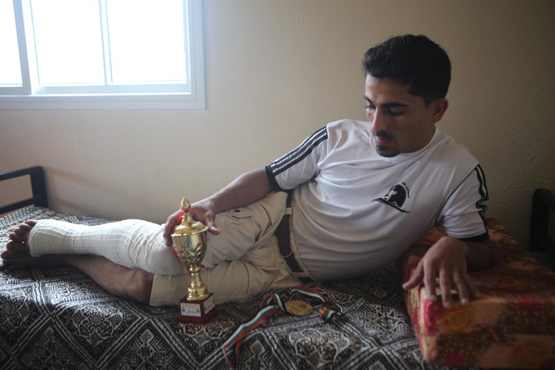 Young man with bandaged leg holds a trophy as he lies across a bed in a sparely furnished room