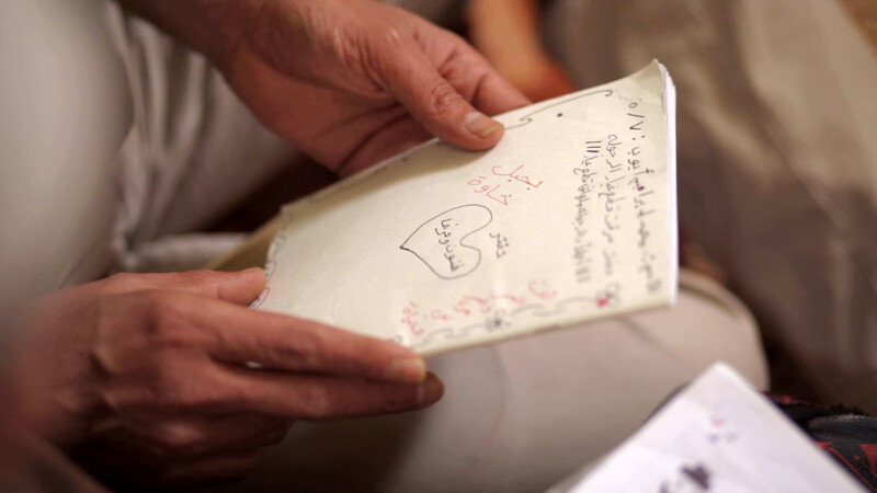 Photo shows two hands holding a notebook with Arabic handwriting on it